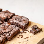 Brownies de chocolate sem ovos com superfície crocante e interior úmido dispostos sobre tábua de madeira ao lado de pedaços de chocolate em barra