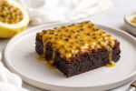 Brownie de maracujá fudgy em prato claro com cobertura amarela cremosa, sementes e fruta ao fundo.