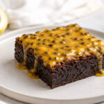 Brownie de maracujá fudgy em prato claro com cobertura amarela cremosa, sementes e fruta ao fundo.