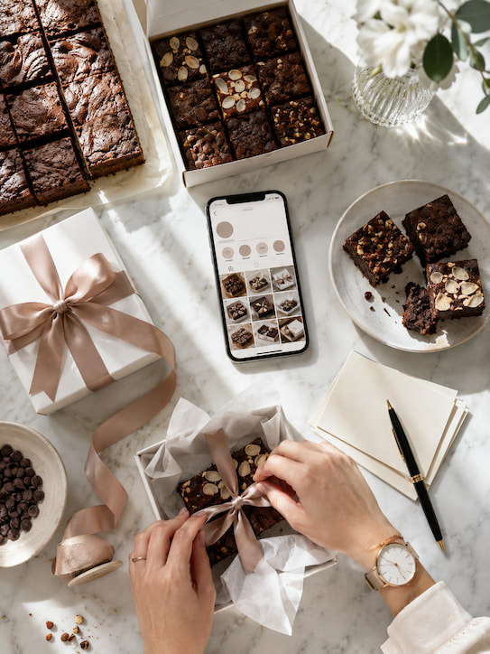 Mesa de mármore com brownies artesanais, celular com destaques do Instagram e caixa de presente sendo fechada.