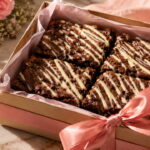 Caixa de brownie para presente de Dia das Mães com laço rosa, cartão carinhoso, flores e caneca ao fundo