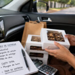 Confeiteira no carro com caixas de brownies, calculadora e caderno sobre taxa de entrega para confeitaria