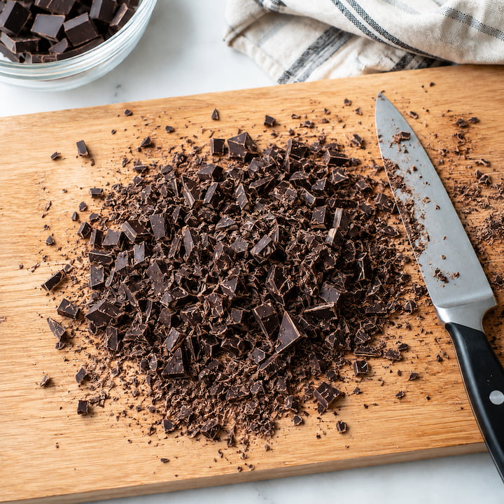 Chocolate picado fino sobre tábua de madeira com faca ao lado, mostrando pedaços pequenos para derreter por igual.