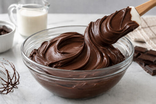 Ganache de chocolate brilhante sendo despejada na tigela, com creme, chocolate e utensílios sobre a bancada.