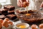 Ovos no brownie é o tema, um ovo aparece sendo quebrado na massa de chocolate, com fouet e pedaços prontos ao lado.