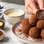 Trufas de chocolate com centro cremoso, cacau em pó e quatro ingredientes da receita profissional na bancada.