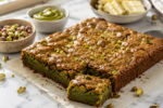 Brownie de pistache com casquinha brilhante e rachada, pistaches picados e fatia cortada sobre bancada clara.