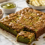 Brownie de pistache com casquinha brilhante e rachada, pistaches picados e fatia cortada sobre bancada clara.