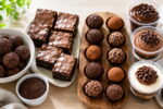 Doces para vender organizados em bancada clara, com brigadeiros, brownies, trufas e bolos no pote em luz matinal.