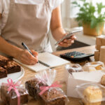 Pessoa organizando renda extra na cozinha com brownies, doces embalados, caixas de entrega e controle de pedidos.