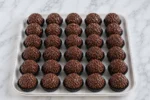 Brigadeiros de chocolate com granulado em forminhas pretas, alinhados sobre bandeja branca em bancada de mármore.