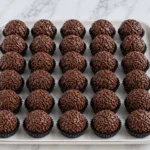 Brigadeiros de chocolate com granulado em forminhas pretas, alinhados sobre bandeja branca em bancada de mármore.