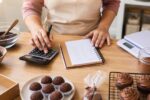 Confeiteira anotando custo de doces com calculadora e caderno; brigadeiros em forminhas e cupcakes na bancada de madeira