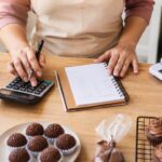 Confeiteira anotando custo de doces com calculadora e caderno; brigadeiros em forminhas e cupcakes na bancada de madeira
