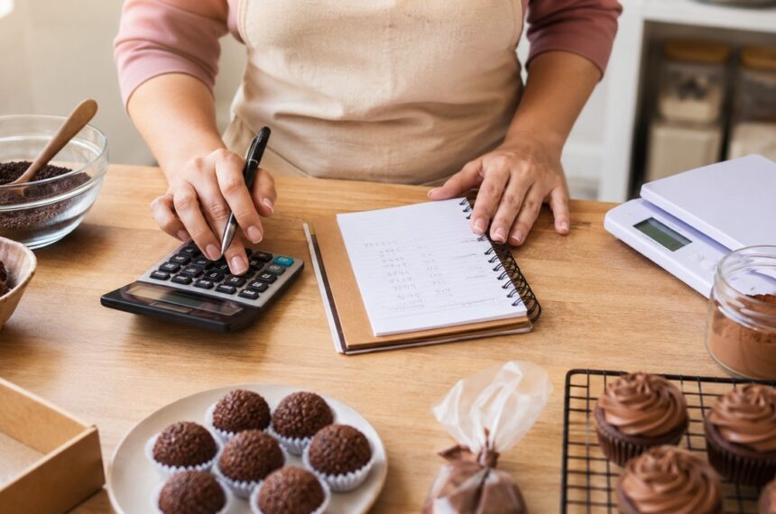 Confeiteira anotando custo de doces com calculadora e caderno; brigadeiros em forminhas e cupcakes na bancada de madeira